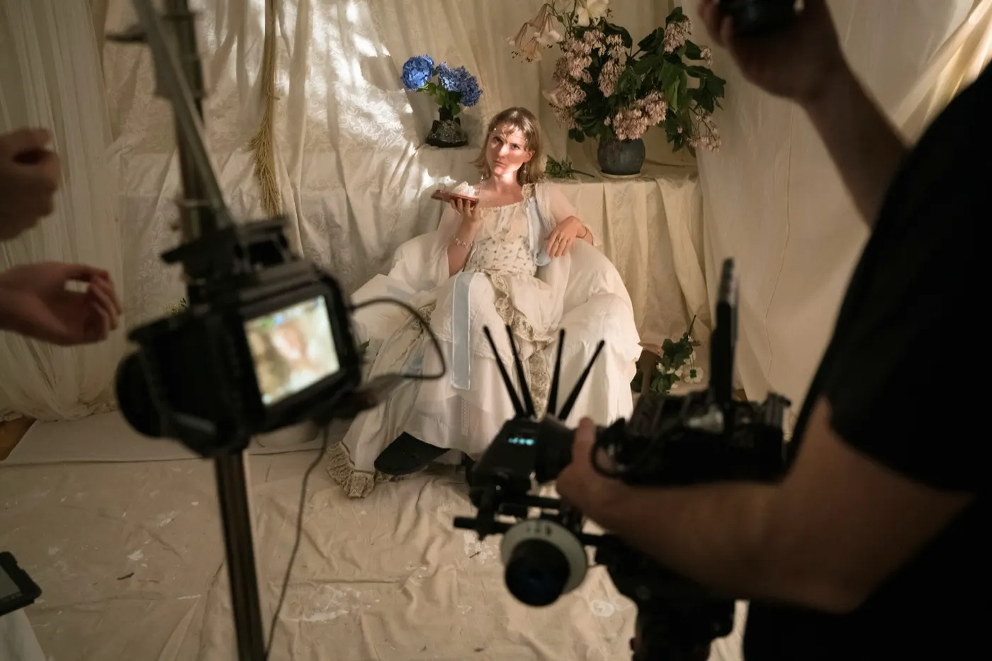 Eva on a music video set, seated in white among flowers, cameras in the foreground
